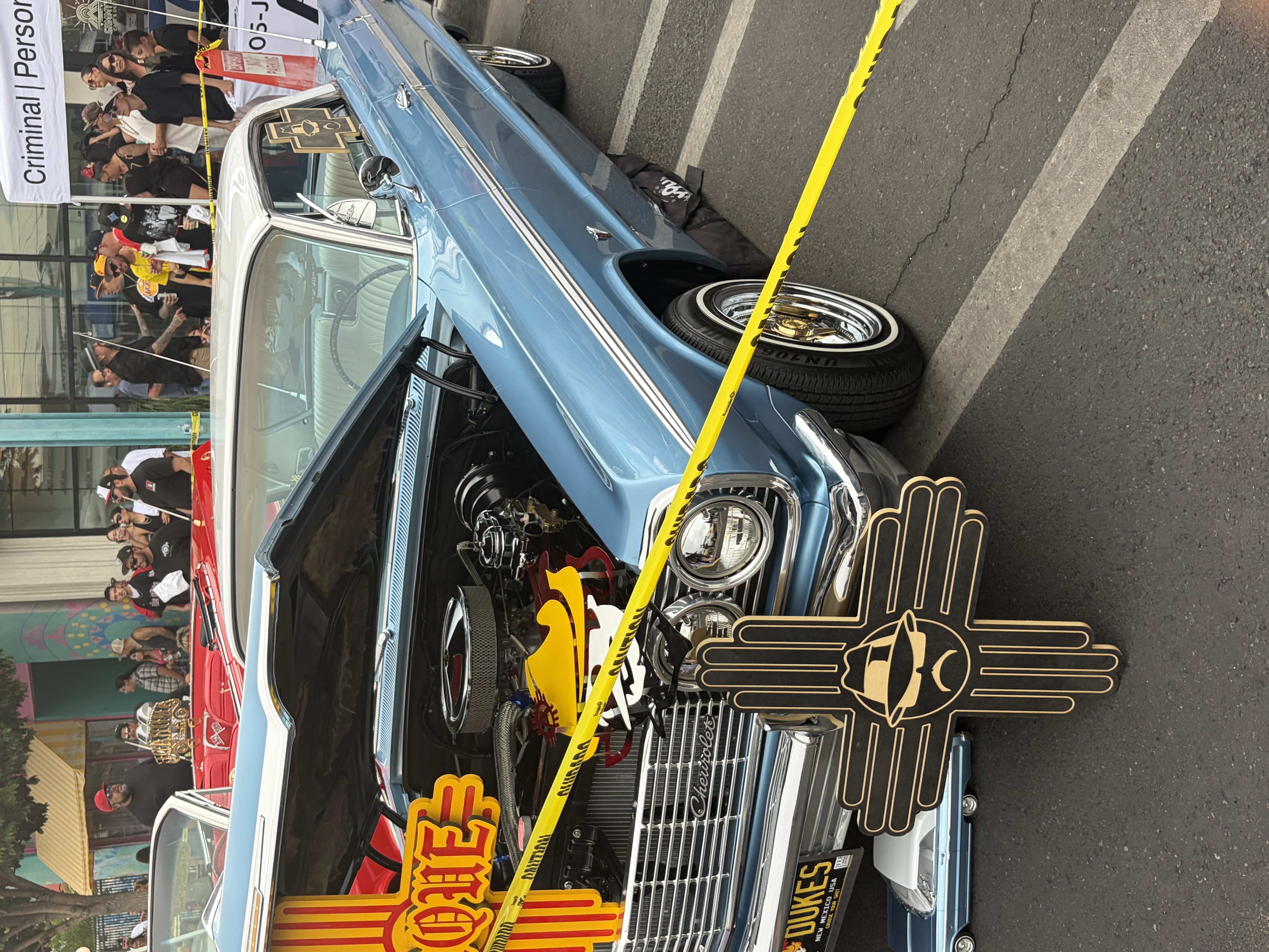 A light blue Impala with the Dukes car club plaque mounted at the front, the Zia symbol above a figure in a hat.